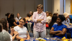 A photo of Gallaudet President Roberta Cordano standing with her arms crossed, looking down attentively to the left at a seated person. The seated person is one of three visible of a group sitting around a circular. The table with has a blue tablecloth with yellow polka dots, and scattered remnants of food and drink. There are other tables and people in the background.