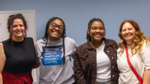 A photo of four people posing and smiling for the camera. They have a variety of outfits and hairstyles. The person second from left wears a gray t-shirt with a logo for the National Museum of African American History & Culture. The background is a light blue wall.