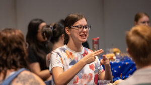 A seated person uses the ASL sign for "name" while smiling at an unseen person to the right. They wear a white t-shirt with colorful pastel flowers, and glasses. In the foreground and background are more seated people.
