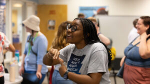 A photo of a standing person making the ASL sign for "hurricane" to an unseen person to the left. They wear a gray t-shirt with a logo for the National Museum of African American History & Culture. In the background, out of focus, are more standing people mingling. They are in a bright room at a school.