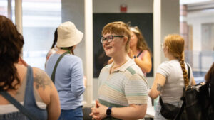 A photo of a person standing and smiling to the left at unseen people. They have short reddish-blonde hair, glasses, and a polo striped in a calm pastel colors. In the foreground and background are more mingling people standing in the bright room.