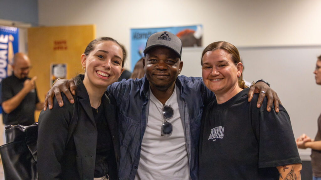 A photo of three people posing and smiling for the camera. The person in the middle has their arms around the other two on the outside. The person in the middle wears a baseball cap, denim jacket, and white t-shirt with sunglasses hung on the neck. They are in a bright room that looks like a school.