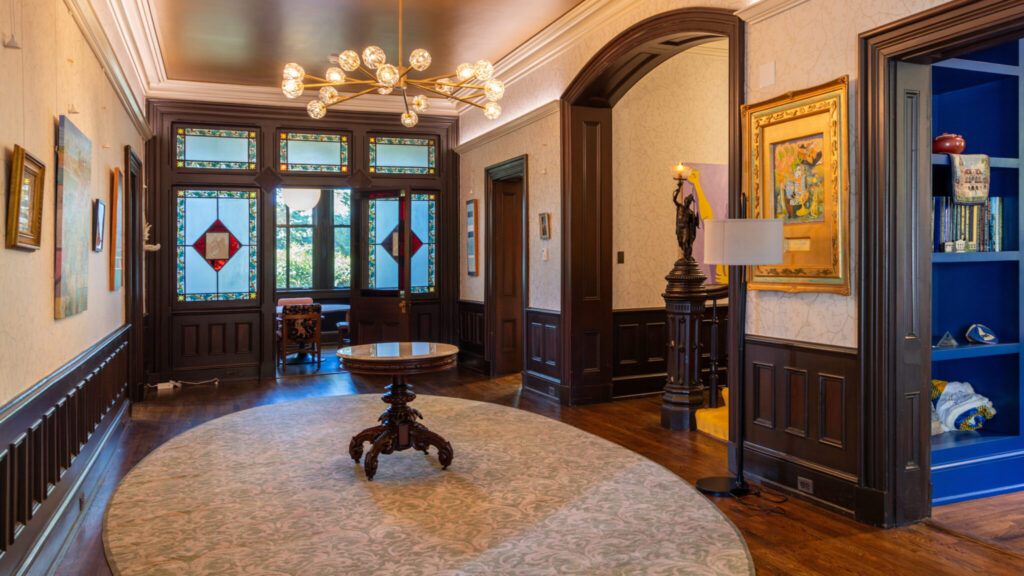 A photo of a hallway inside of a Victorian-style house. The walls and doorways have deep brown wooden fixtures and beige wallpaper. Notable features include golden-framed art, a small circular table in the center of the walkway, and an entrance with stained-glass door windows in the back.
