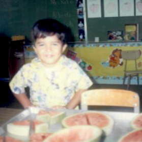 An old photo of a child in a classroom, smiling for the camera. The child has short dark hair. In the foreground is a table with slices of watermelon.