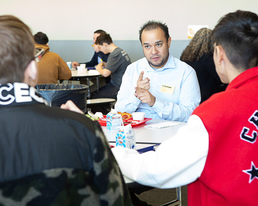 A photo of a person sitting at a table, signing sincerely towards two youth in the foreground. The person has soft, spiky dark hair. They wear a light blue button-up and a "Visitor" tag. In the background are more young people sitting around tables.