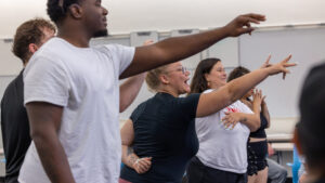 A photo from the side of five people signing with arms outstretched to the right. In the foreground on the left, a student wears a white t-shirt. In the middle, a student wears a black t-shirt and their mouth is open with enthusiasm.