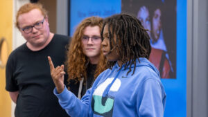 A photo of three students student at the front of a classroom. In focus in the middle is a student with a light blue hoodie that has a large "C" and small "Chicago". They are making a gesture that looks like the ASL sign for the number one. Behind, two out-of-focus students watch.