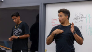 A photo of a student standing at the front of a classroom, in front of a whiteboard. They are making a gesture that could be a number of different signs, with their left hand raised and right hand pointed to their chest. They wear a black t-shirt and gray pants. Two other students are out of focus on the left.