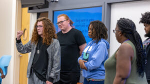 A photo of five people standing at the front of a classroom. In the front is a student making the ASL sign for "A". They wear a stylized monochrome jacket, black shirt, dog tag necklace, and gray pants. Around them, the other four people watch.