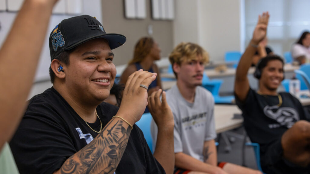 A photo of a seated smiling student making the sign for "C". They wear a black hat and t-shirt. They have a pinky ring, gold bracelet, and forearm tattoo. A blue earmold is visible in their ear. In the foreground on the left a raised arm is partially visible. In the background to the right are out-of-focus students.