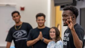 A photo of four students standing at the front of a class. Only the student on the right is in focus. They smile while making the ASL sign for "D" with their left hand. They wear a black t-shirt and have black shair with a light beard.