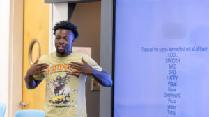 A photo of a student signing in front of a classroom digital screen. The student has tall curly hair with shaved sides, and a goatee. They wear a colorful green shirt and camo pants. The screen says "These all the signs i learned but not all of them", followed by a list of words.