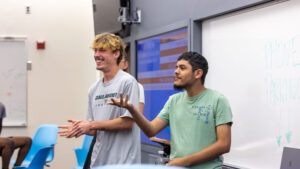 A photo of two smiling students in front of a classroom. The student on the right is making a thoughtful gesture toward the audience offscreen to the left. The student on the left is amused.