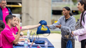 A photo of an outdoor reception desk during a college move-in day. Two standing students on the right smile at seated workers on the left wearing pink shirts. Behind the foreground, a worker is handing a bag that says "Gallaudet University" to a student.
