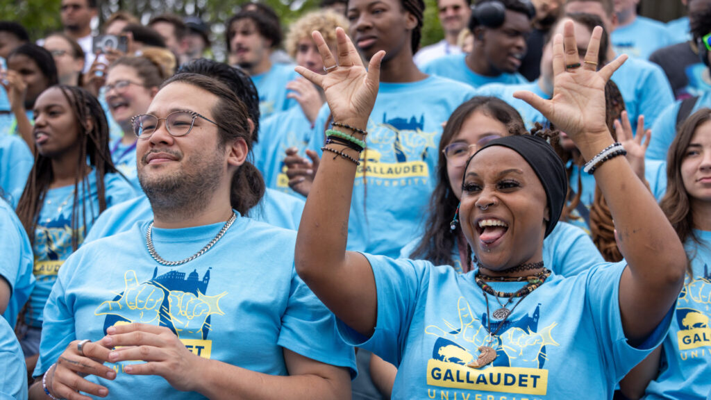 A photo of a crowd of people in bleachers. Everybody is wearing the same light blue t-shirt, with "Gallaudet University" and an image of hands making the ASL sign for Gallaudet. In the foreground, a person on the right raises their hands in excitement. They have a variety of accessories and piercings. On the left, a person smiles softly.