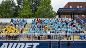 A photo of dozens of new college students sitting on football bleachers, smiling for the camera. On the right, the majority of the group is wearing light blue t-shirts. Some have yellow shirts, including another group on the left across the aisle. In the front Gallaudet President Roberta Cordano stands between students. Behind the bleachers, a group of trees is visible in the top left, and a building is visible in the top right.