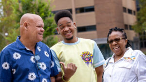 A photo of a family laughing together. In the center is a student with a colorful yellow t-shirt, hi-top haircut, chin beard, and hearing aids. The student is pointing their right thumb to the older, bald, masculine-presenting person on the left of the image, who is wearing a blue shirt with a flower pattern. On the right of the image is a shorter, feminine-presenting person with black hair, sunglasses, and a white shirt.