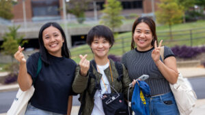 Three students smile for the camera. The two students on the left make finger hearts with their right hand thumb and index fingers. The student on the right makes the peace sign with their left hand. In the background is the Gallaudet campus, with a road, greenery, and building.