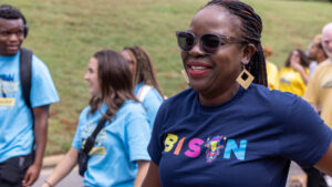 A photo of people in colorful t-shirts walking on a road. Prominent in the foreground on the right is Gallaudet Provost Khadijat Rhashid, smiling and walking to the left. Provost Rhashid has long hair in braids, tied back in a ponytail. She wears sunglasses and a dark blue shirt that says "Bison" in colorful text.