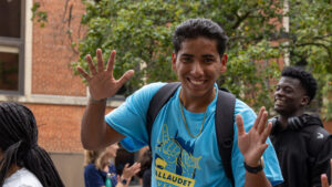 A photo of a student smiling broadly and making handwaves toward the camera. They have short, poofy black hair. They wear a light-blue t-shirt that says "Gallaudet", a gold chain, watch, and black backpack. Behind and out-of-focus are more smiling students. In the background are a tree and building.
