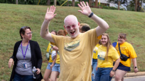 A photo of an excited person smiling broadly and raising their hands in an enthusiastic greeting toward the camera. They have a close-trimmed beard. They wear a yellow t-shirt, white-rimmed sunglasses, and watch. Behind and out-of-focus are more smiling students in yellow t-shirts on the right, and a middle-aged person on the left. In the background is a grassy knoll.