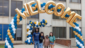 A photo of four smiling people standing under a large arch that is wrapped in blue, white, and gold balloons, and fronted on top with separate shiny golden balloons that spell out "Welcome". The person on the left is the tallest and likely the oldest, while still young. The three on the right are younger, in a variety of colorful outfits. In the background is a concrete building with more balloons around a doorway.