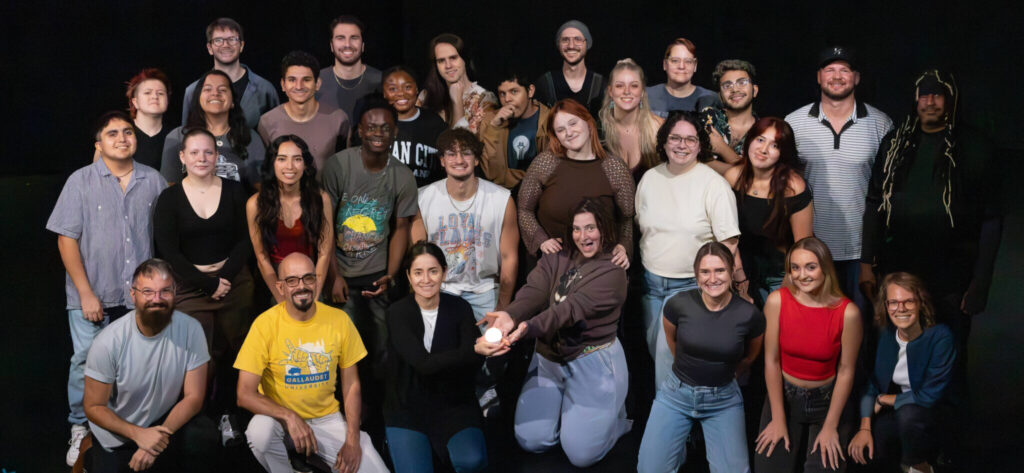 A large group of diverse individuals, approximately 40 in total, posing together in a theater setting. They are arranged in a semi-circle, with a mix of genders and ethnicities. Some are smiling, and they are dressed in casual clothing. The background is dark, emphasizing the group in the foreground.