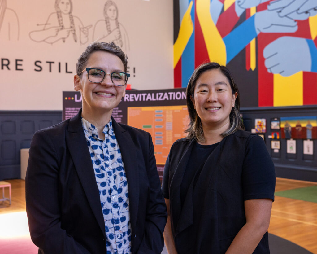 A photo of two people standing and smiling in a museum exhibit. The person on the left has short gray hair, glasses, a dark blazer, and a patterned button-up. The person on the right is slightly shorter. They have shoulder-length dark hair and a dark shirt. The museum in the background has a colorful mural visible in the top right.
