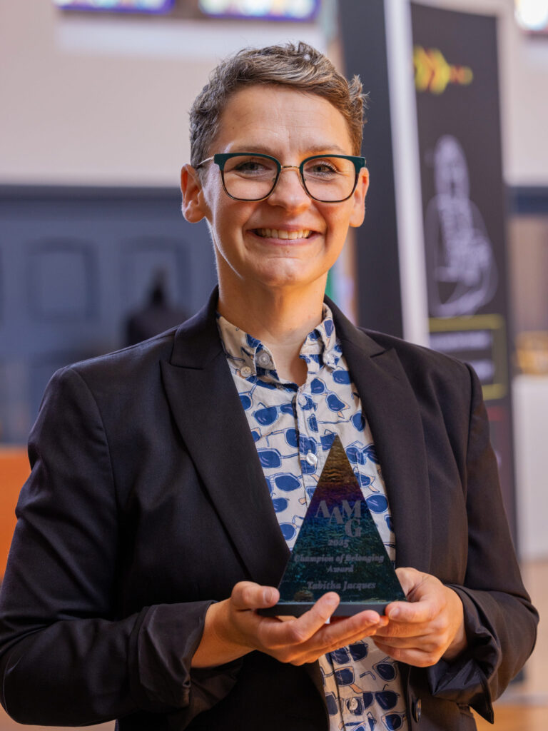 A photo of Tabitha Jacques posing with an award. Tabitha has short gray hair, glasses, a dark blazer, and a patterned shirt. The award is a dark, pyramid-shaped piece of stone.