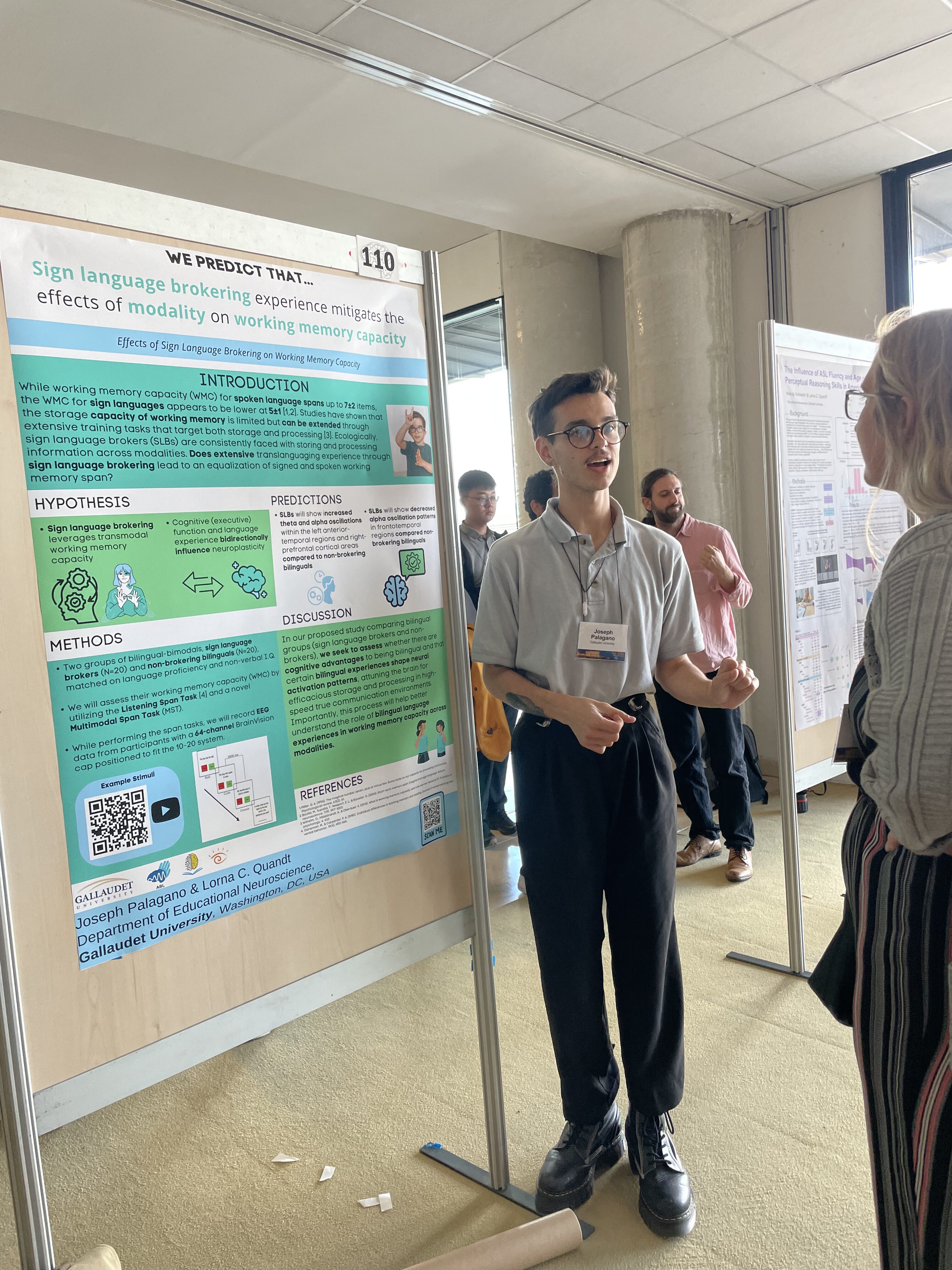 Man stands next to a scientific poster at an event. He appears to be explaining some information to a person standing in front of him. There are other posters on bulletin boards and other people in the background.