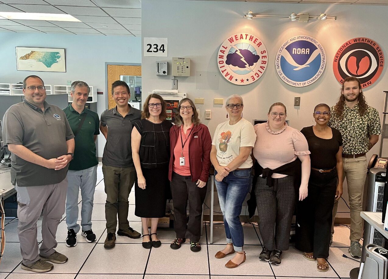 Nine people stand in an office setting. Behind them is a wall with circular signs. One reads, "National Weather Service" and depicts a dark cloud with a lightning bolt in the middle. Another has the symbol of NOAA.