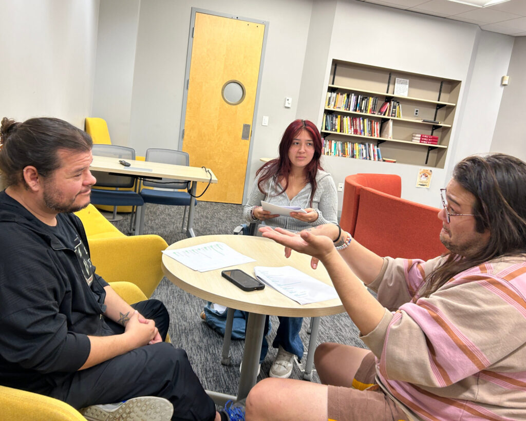 Three university students sit around a table practicing a performance. Each student has a paper script. The student on the bottom right side of the table is making a dramatic gesture. The student in back watches attentively. A door and built-in bookshelf are visible in the background.