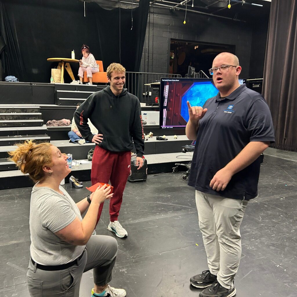 Three university students rehearse in a black box theatre. In the foreground, the student on the left is kneeling with a box and smiling at another student on the right. The student on the right is looking offscreen. The student in the back looks on with a smile. In the background are rows for seating.