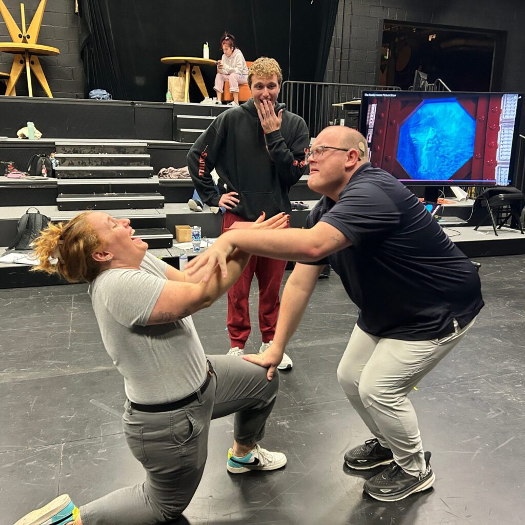 Three university students rehearse in a black box theatre. In the foreground, the student on the left is kneeling with a box and reeling away from the student on the right, who is dramatically leaning on them. The student in the back looks on with a smirking surprise. In the background are rows for seating.
