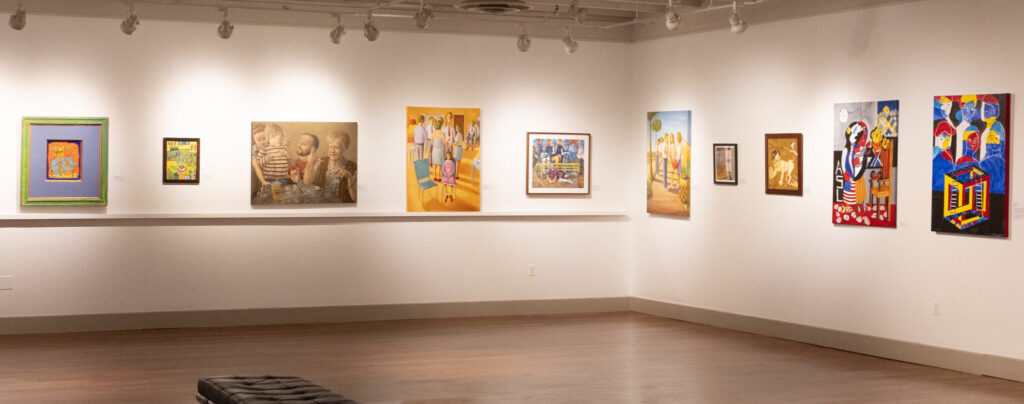 A wide photo of art on the wall of a gallery. The art varies in size, color, and subject, clearly by different artists. The gallery is otherwise nondescript, with pale walls and a wood floor.