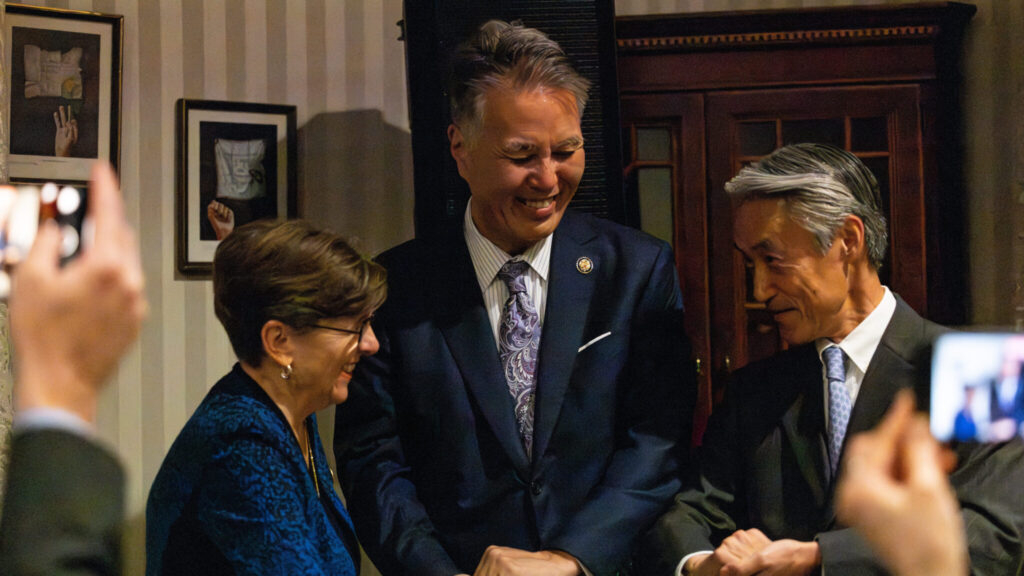 Three well-dressed, dignified people smile warmly at each other. The person on the left is shorter, with short brown hair, glasses, earrings, and a necklace. They wear a strikingly patterned blue and black jacket over a black shirt. The person in the middle has kempt black and silver hair. They wear a well-fitting blue suit. The person on the right has coifed silver and black hair. They wear a fitted gray pinstriped suit with a white shirt and blue tie. The background is a stylish home.