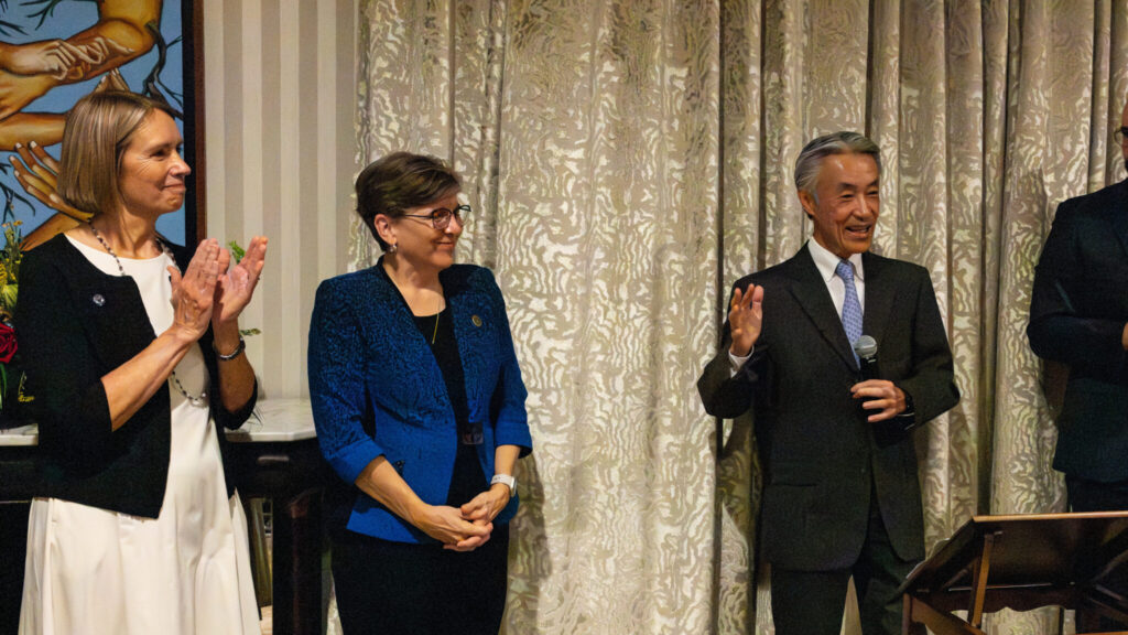 A person in a grey suit, center right, holds a microphone and speaks to an unseen audience on the right. They have coifed silver and black hair. On the left, two well-dressed people stand smiling. The person on the far left is applauding.