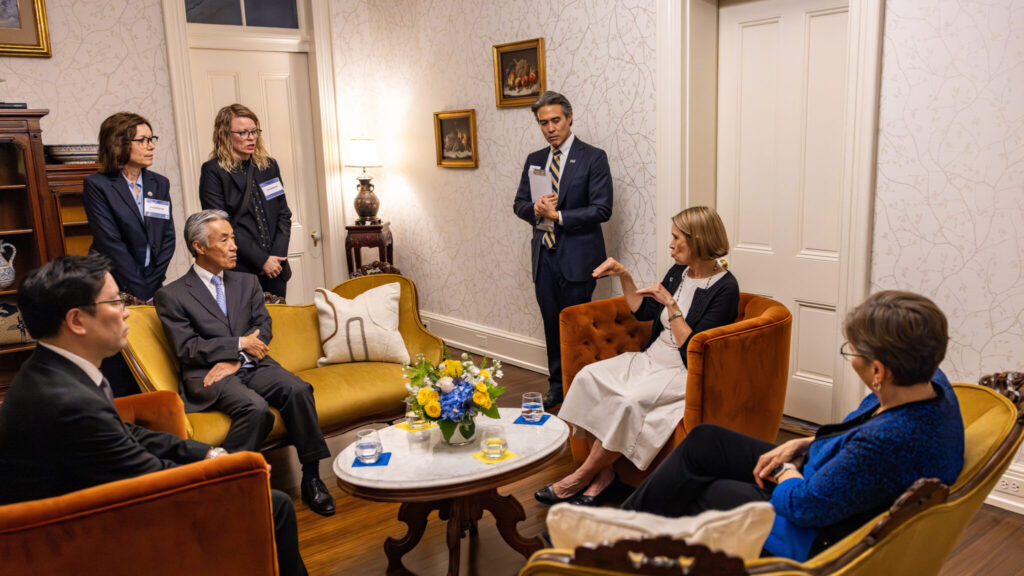 A photo of seven well-dressed people in a room with upscale decor. Three are standing and four are sitting. Most are looking at a seated person, second from the right, using sign language. The person to their right has a clipboard.