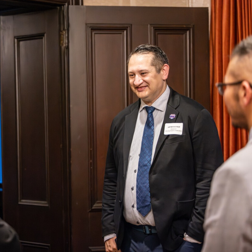 A person in a dark suit stands and smiles to somebody offscreen to the left. They have slick brown hair.