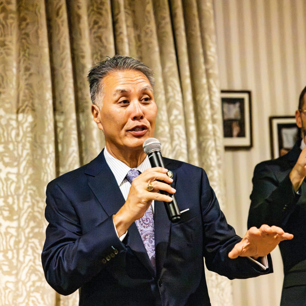 A person in a well-fitting blue suit speaks into a microphone for an unseen audience to the right. They have kempt black and silver hair and are making a dignified gesture to the audience. A sign language interpreter is partially visible in the background on the right.
