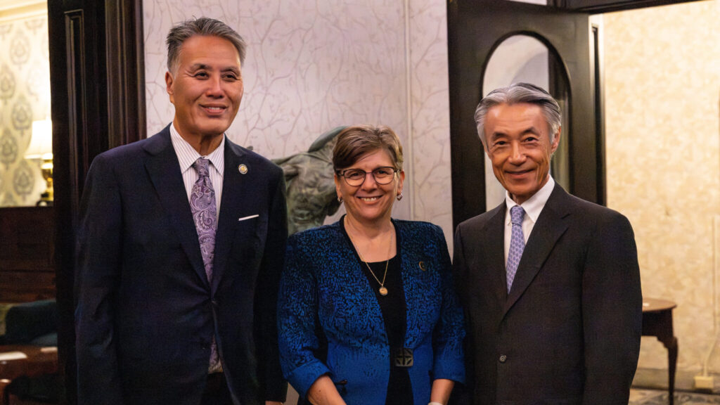 Three well-dressed, dignified people smile for the camera. The person on the left has kempt black and silver hair. They wear a well-fitting blue suit. The person in the middle has short brown hair, glasses, earrings, and a necklace. They wear a strikingly patterned blue and black jacket over a black shirt. The person on the right has coifed silver and black hair. They wear a fitted gray pinstriped suit with a white shirt and blue tie. The background is a stylish home.