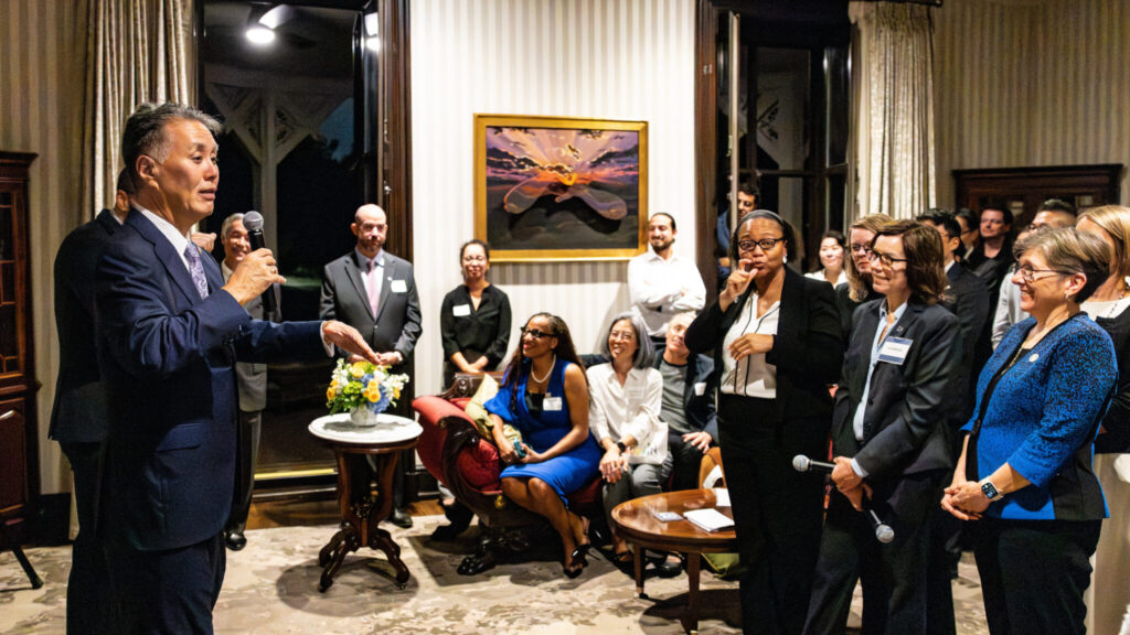 A photo of a person, on the left, speaking into a microphone to an audience in a small room with upscale decor. The person speaking is wearing a blue suit and gesturing. The audience well-dressed, and most are smiling at the speaker.