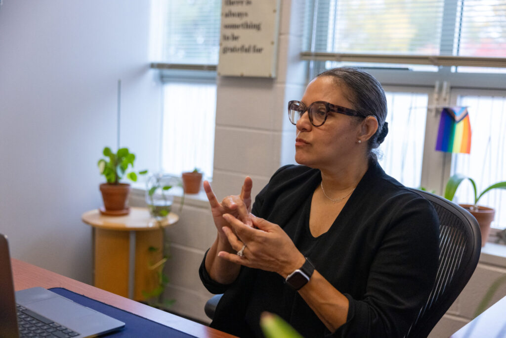 A woman with glasses is seated at a desk, using sign language while engaging in conversation. She is wearing a black top and appears focused. In the background, there are potted plants and a rainbow flag.
