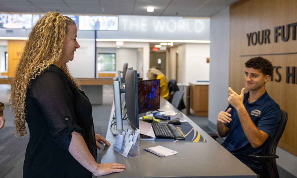 A woman with long, curly hair is standing at a reception desk, engaging with a young man who is seated behind the desk. The man is using sign language while communicating. There are computers and office supplies on the desk, and the background features a modern office environment with signage that reads "YOUR FUTURE STARTS HERE."