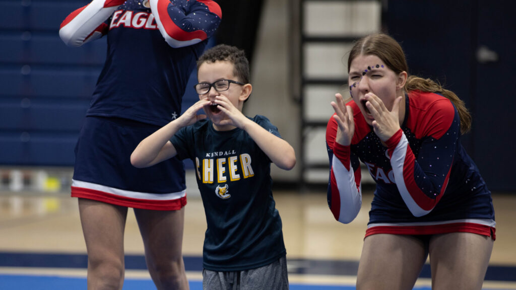 A photo of two cheerleaders standing on either side of a child. The cheerleader on the left is only visible from the chest down. The child and cheerleader on the right are leaning over and cheering enthusiastically. The child's dark blue shirt shirt reads, "Kendall Cheer". The cheerleader's uniforms are dark blue and red.