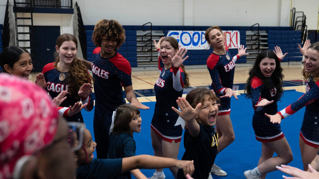 A photo of ten people, including at least six cheerleaders in uniform, smiling and cheering in a school gym. The cheerleaders are standing around three children in front, making various hand gestures that evoke cheer.