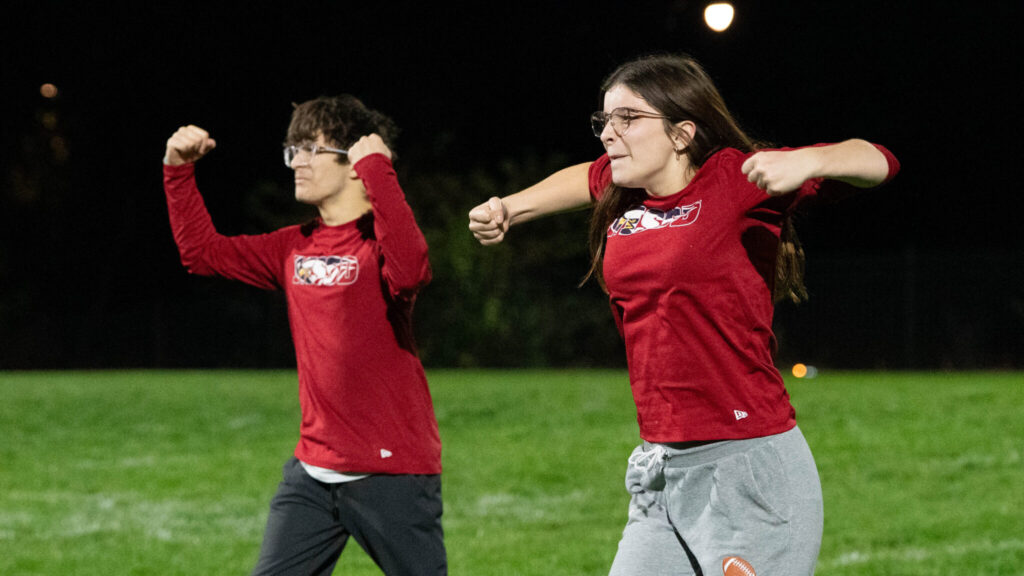A photo of two young people with their arms raised to their chest in the midst of a dance on a football field at night. They look intensely at an unseen crowd to the left. Both wear long-sleeved red shirts with an "MSSD" logo.