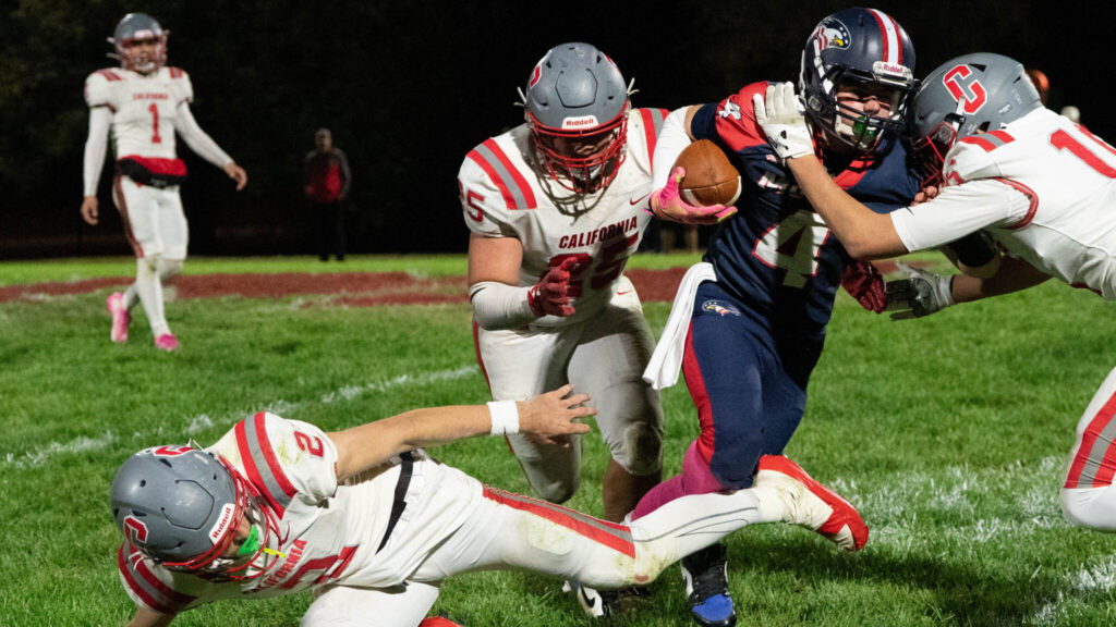 A photo of five young football players during a game. Four have white uniforms, and one has dark blue. In the foreground, the dark blue player attempts to run with the ball, with two white-uniformed players in front and behind. Another white-uniformed player is down on the ground. In the background, a white-uniformed player watches.