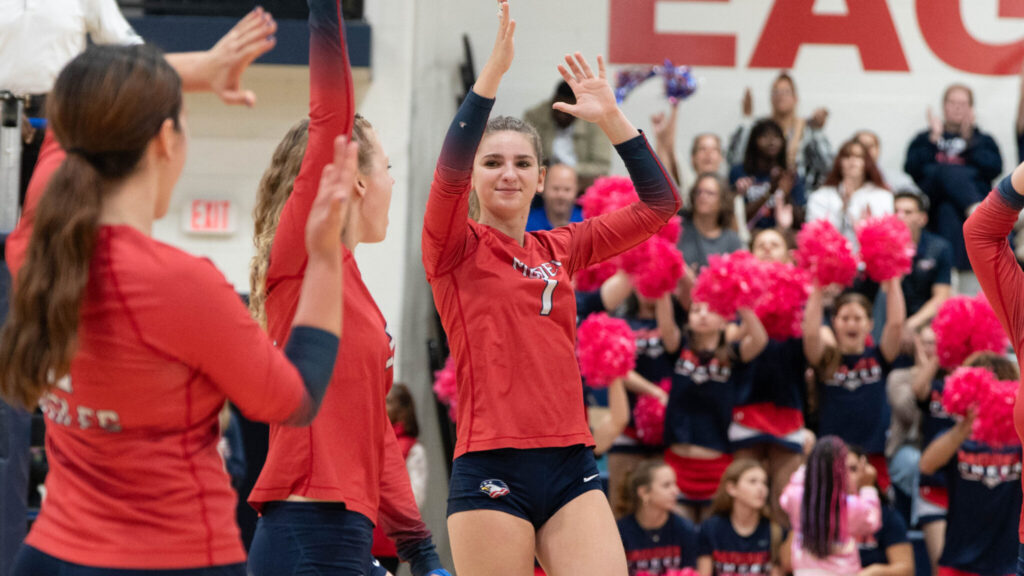A photo of three young volleyball players, with a crowd on bleachers in the background. The players are wearing red long-sleeved shirts and black shorts. Prominent in the middle, facing the camera, a player smiles and raises their hands in celebration. The crowd in back includes several people raising red pompoms.