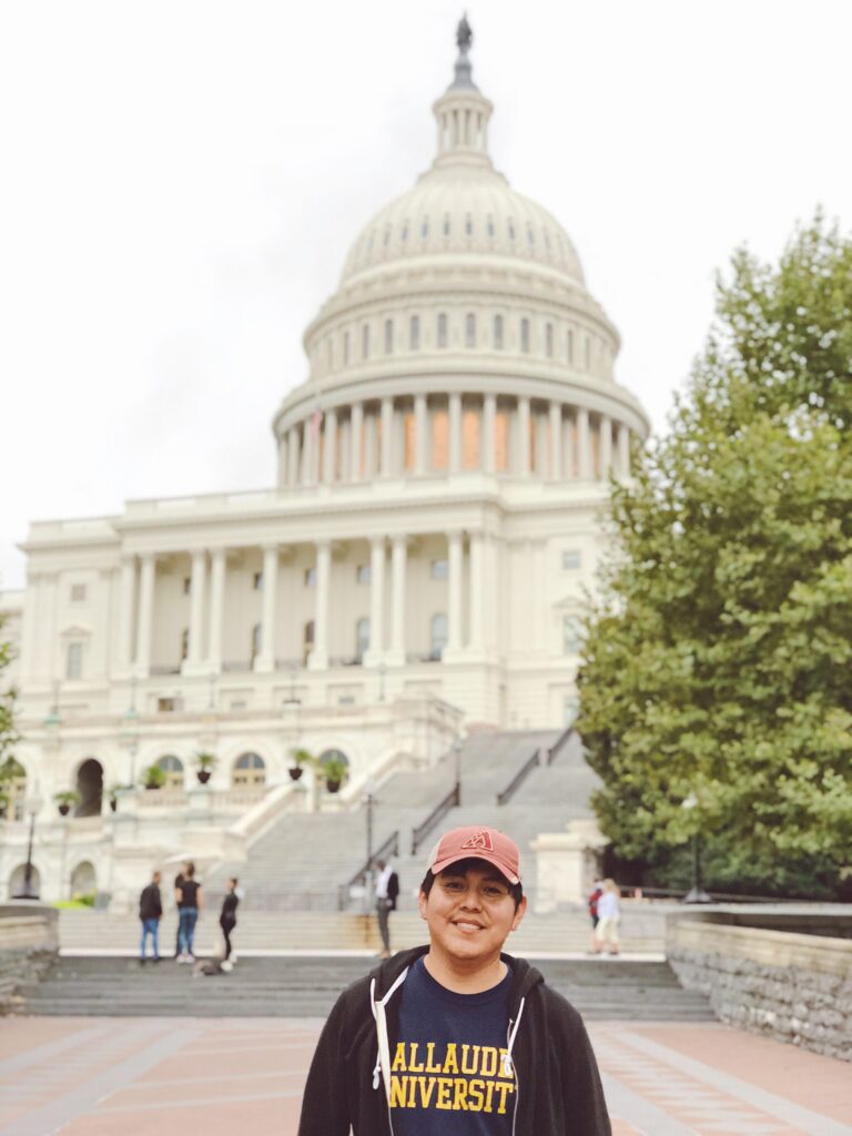 A photo of a student posing in front of the US Capitol Building. They wear a cap and Gallaudet University shirt.